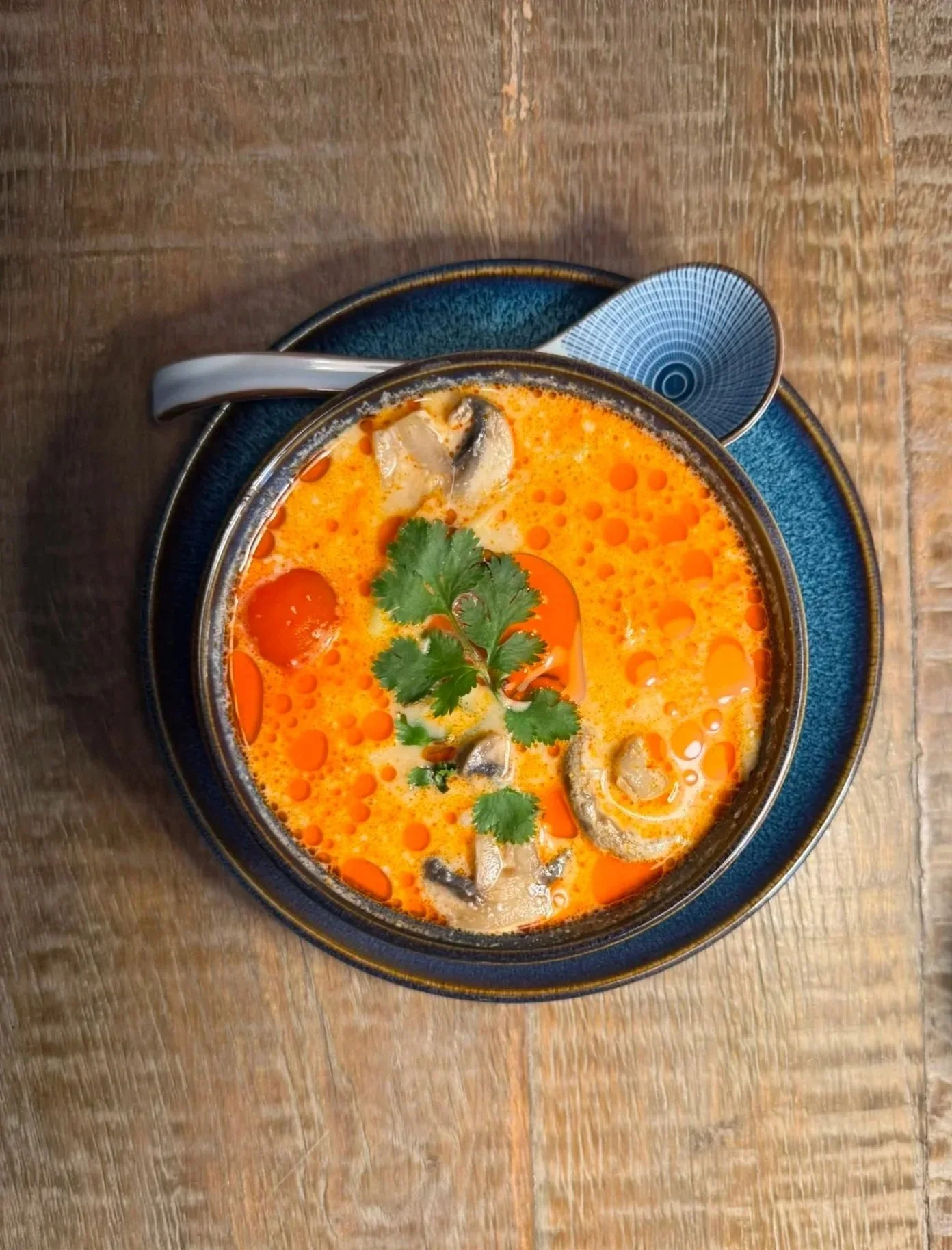 A bowl of orange-colored soup with cilantro, sliced vegetables, and herbs, served on a blue plate with a spoon, on a wooden table.