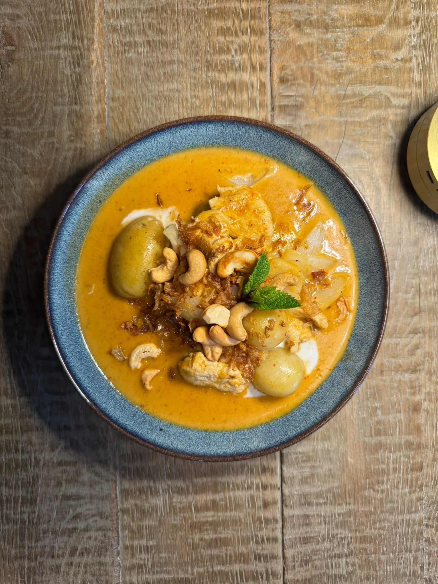 A bowl of yellow curry with potatoes, chicken, cashews, and vegetables garnished with a green mint leaf, placed on a wooden table.