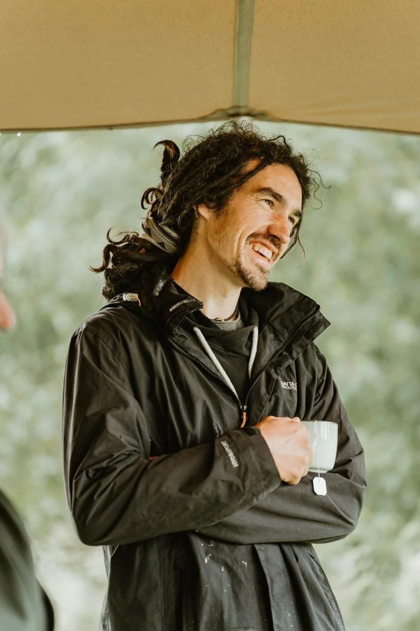 A man with long dreadlocks, a beard, and wearing a black jacket is holding a white mug and smiling, standing outdoors under a canopy with green trees in the background.