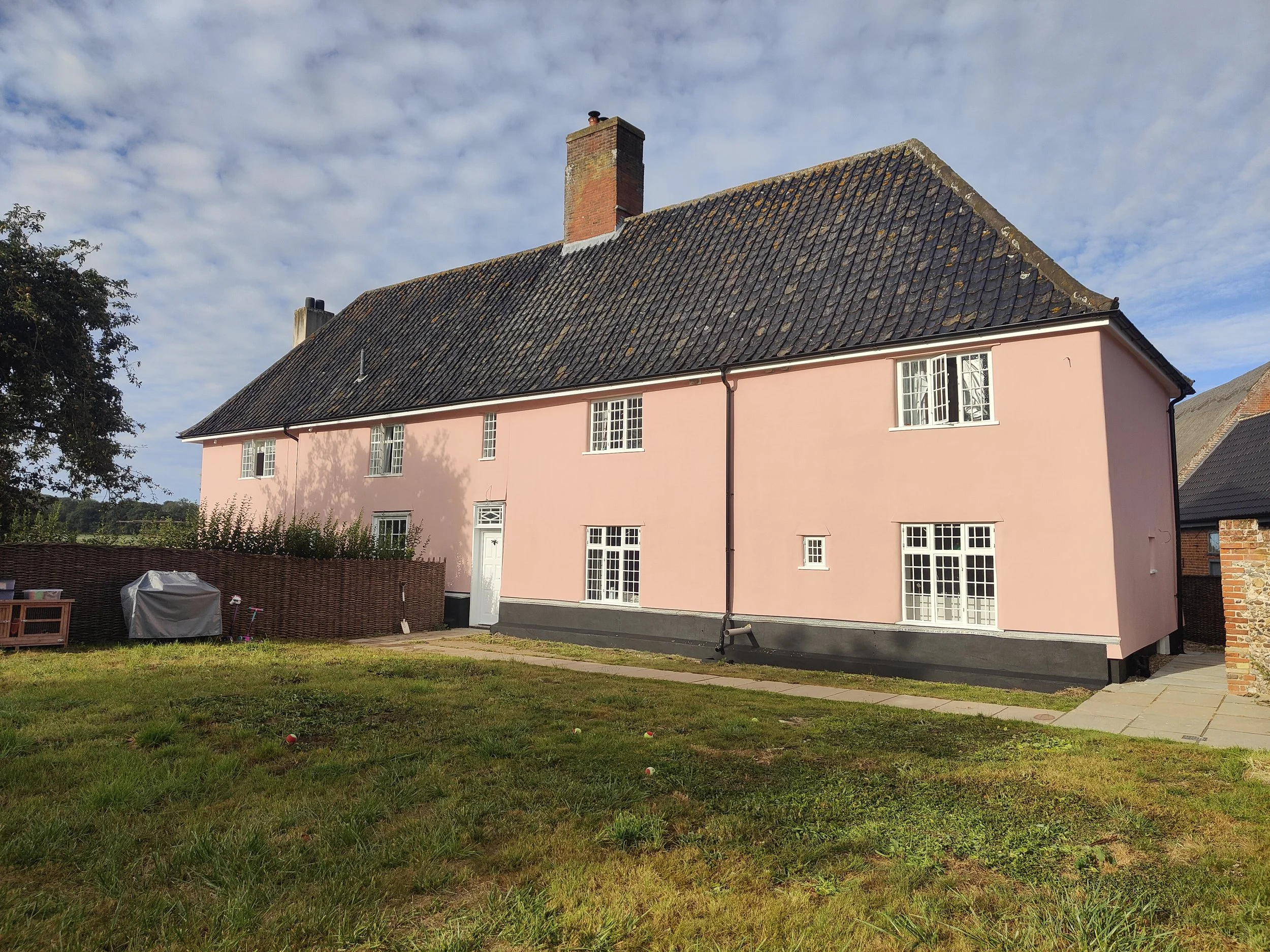 A two-story house painted pink with white-framed windows and a black roof, situated on a grassy yard with a paved pathway. The sky is partly cloudy, and there is a brick wall and outdoor furniture or storage covered with a tarp on the left side.