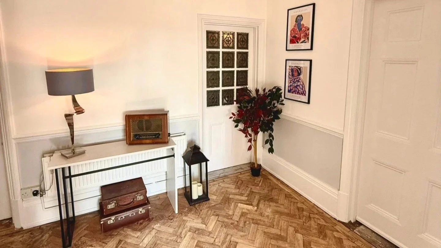 A corner of a room with a white wall, hardwood herringbone floor, a white console table, a modern table lamp, a vintage radio, a lantern with candles, a potted plant, framed artwork on the wall, a door with glass blocks, and a closed white door.