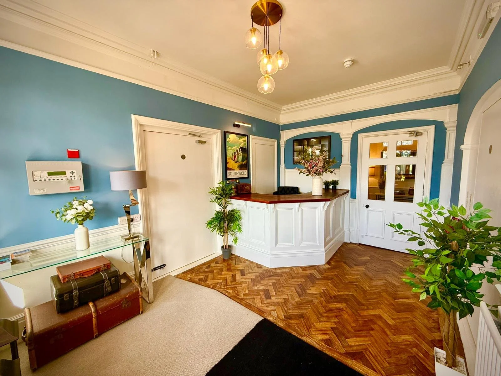 Hotel reception area with blue walls, wooden flooring, white decorative molding, a white front desk with a floral arrangement, potted plants, and a wall-mounted art piece.