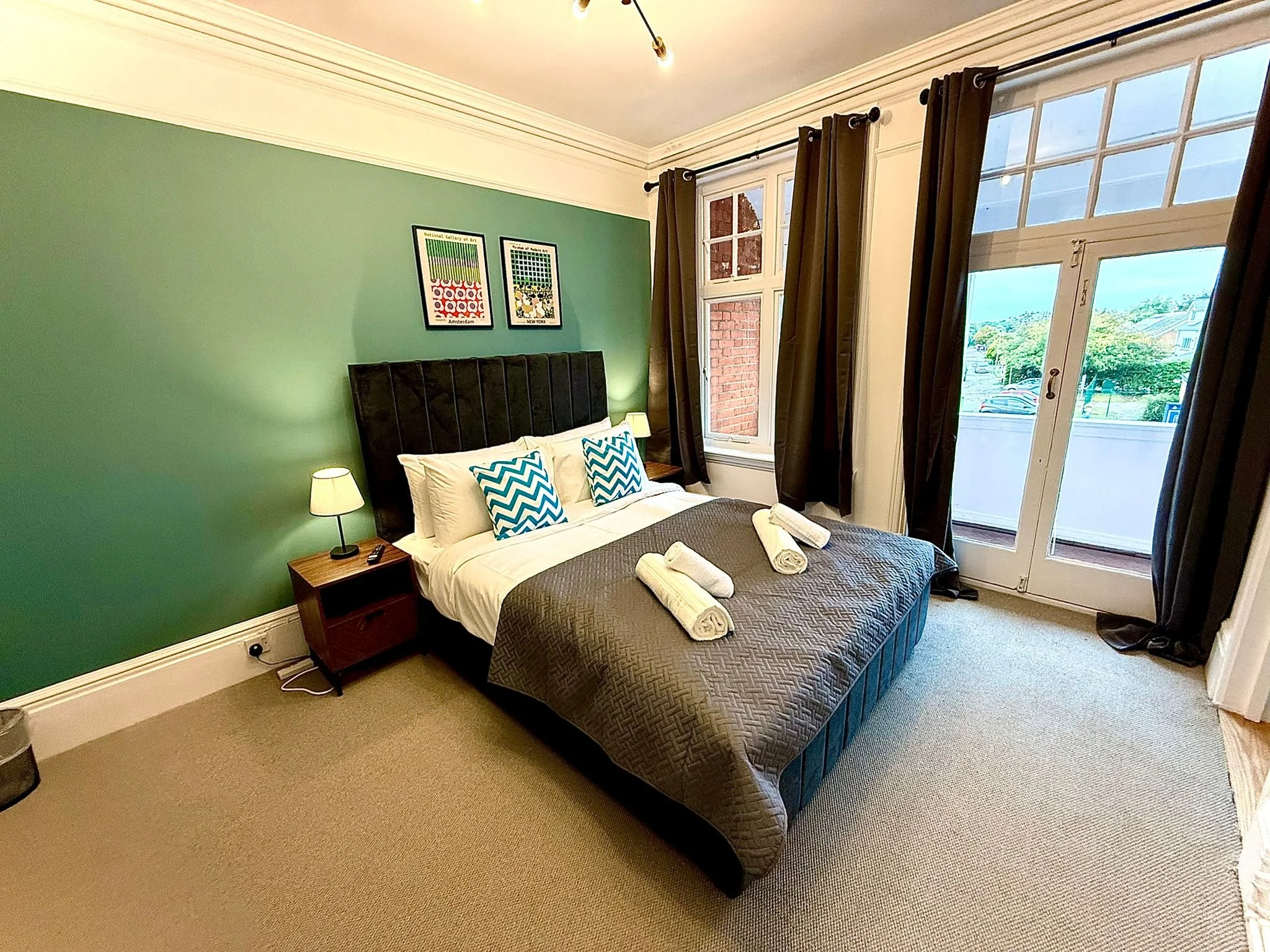 Bright bedroom with green accent wall, large bed with white pillows, blue and white chevron pillows, dark gray quilt, and three rolled towels. Two nightstands with lamps, a window with dark curtains, and a glass door leading to a balcony with a view of trees and parked cars.