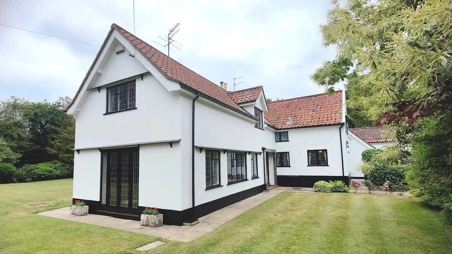 A white two-story house with a red tile roof, surrounded by a lawn with small bushes and trees.