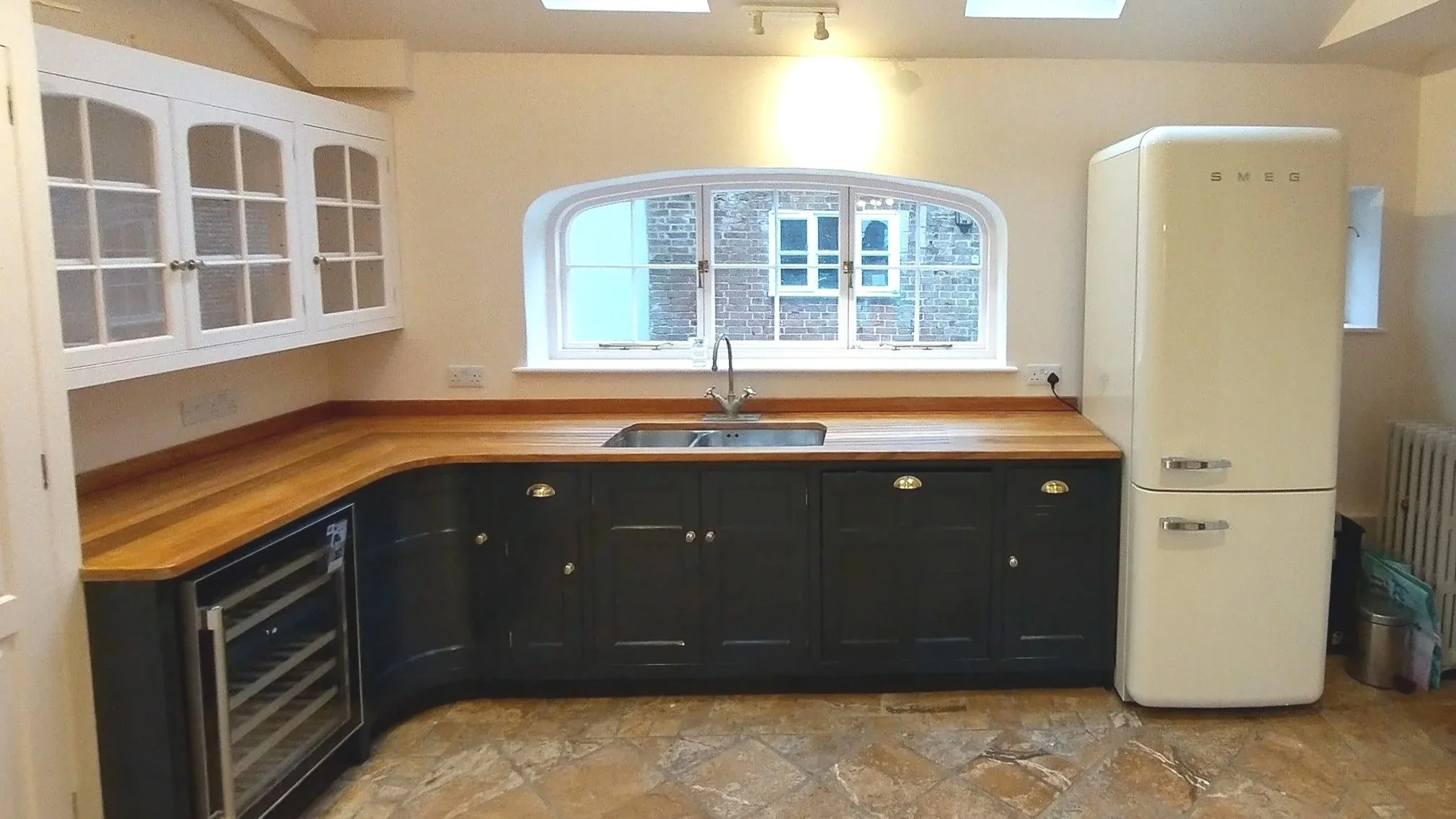 Kitchen with black cabinets, wooden countertop, window above sink, cream SMEG refrigerator, wine cooler, and brick building visible through the window.