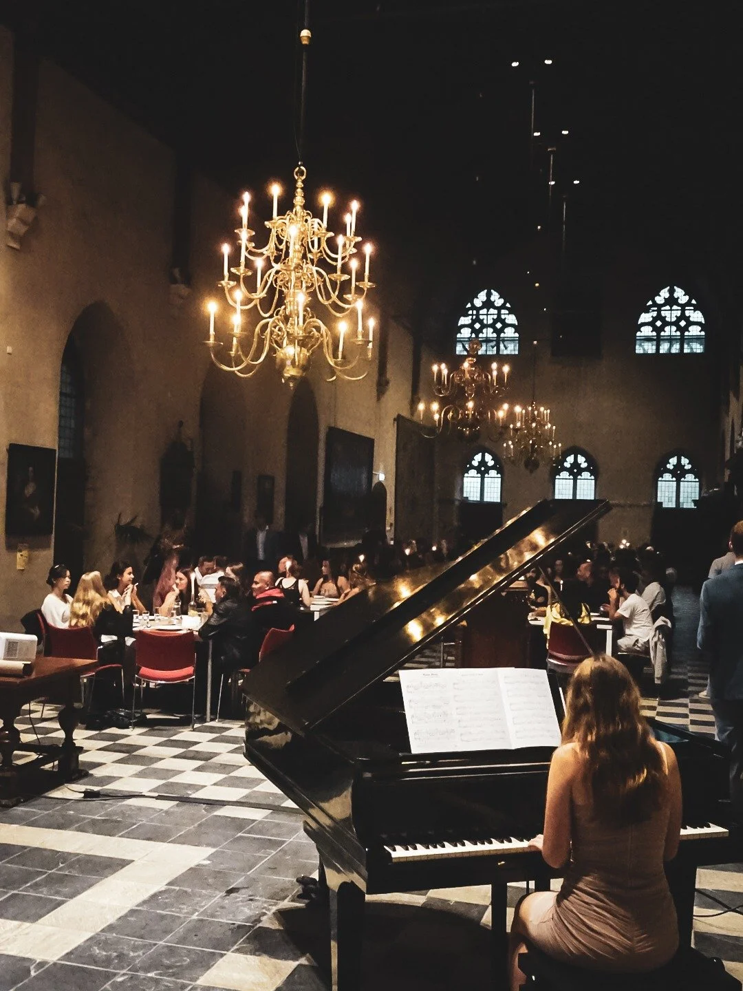 Binnen in een kerk of oude zaal, een vrouw speelt piano voor een publiek, met grote kroonluchters en hoge, gebrandschilderde ramen op de achtergrond.