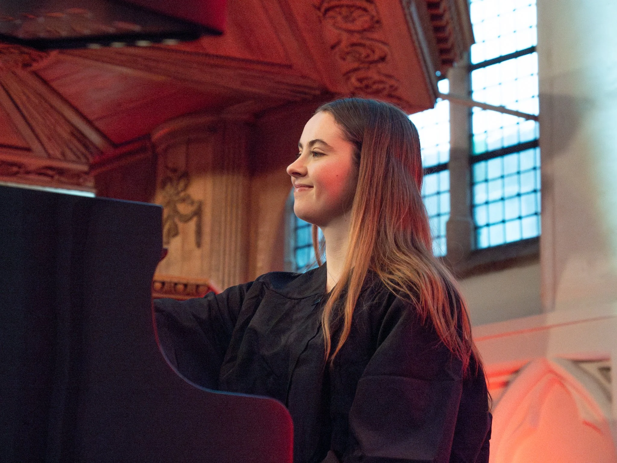 Een jonge vrouw met bruin haar die een piano bespeelt in een grote, met hout beklede zaal met grote ramen en decoratieve versieringen aan het plafond.