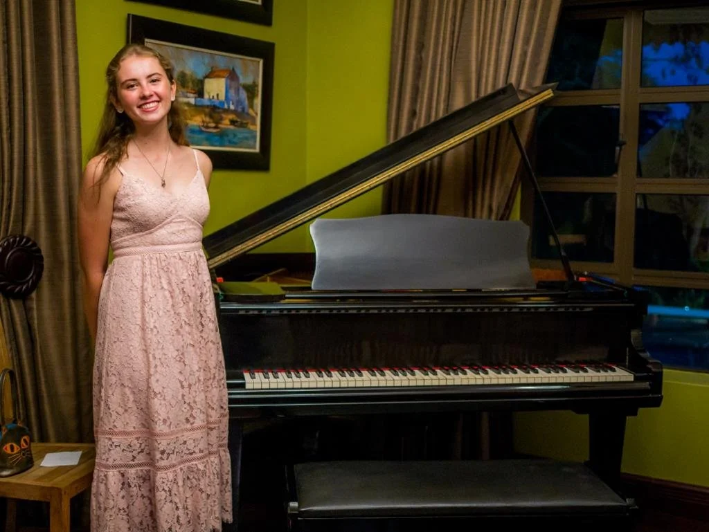 Vrouw in een lichtroze kanten jurk staat naast een piano in een kamer met groen en geel gekleurde muren, schilderijen en een raam.