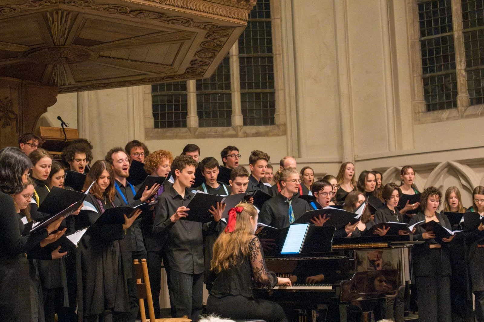 Koorzang in een kerk met hoge ramen, met een dirigent die voor de groep zit, en een piano erbij.