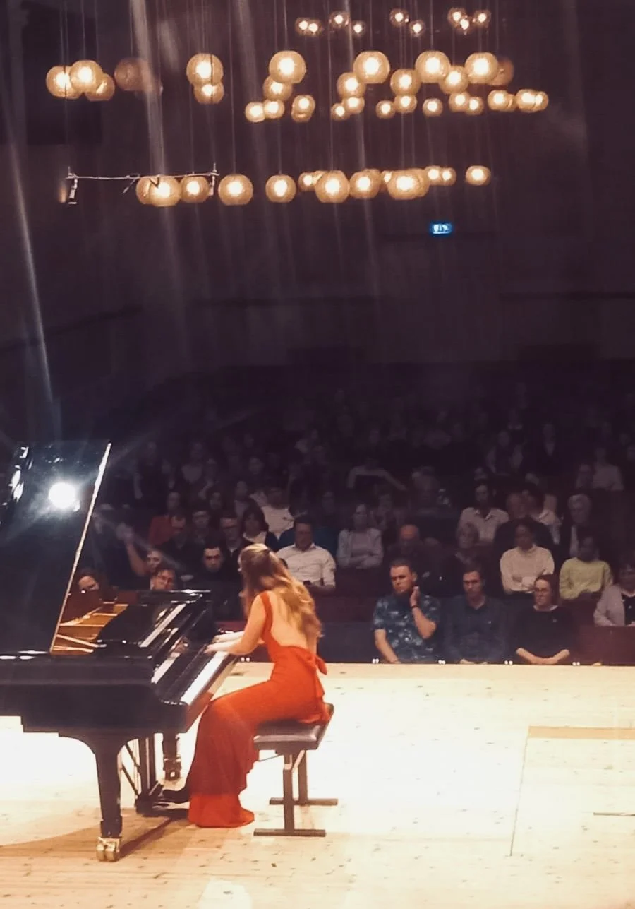 Een vrouw in een rode jurk speelt piano op een podium voor een publiek in een concertzaal.