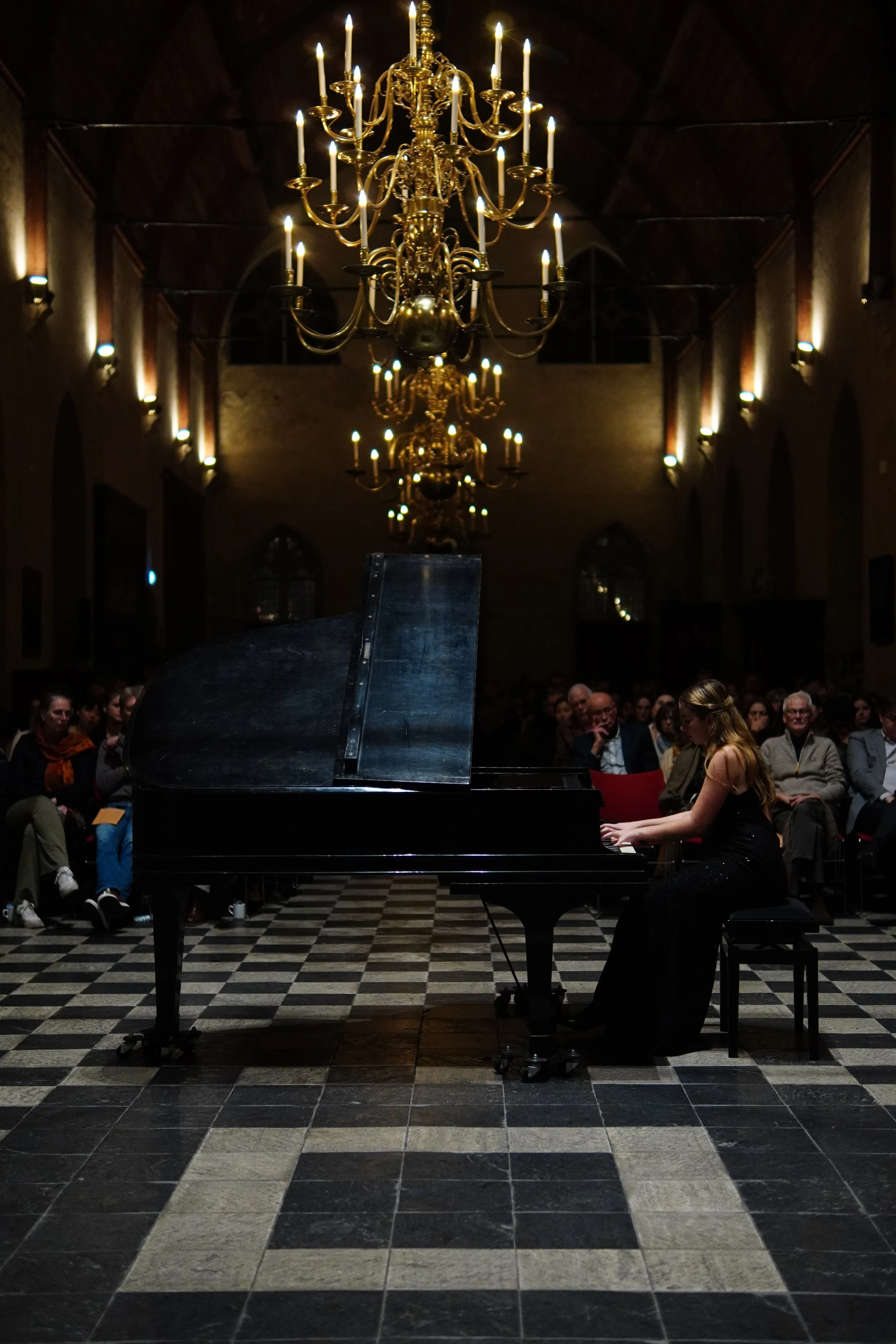 Een muzikant speelt piano tijdens een concert in een grote zaal met een chique kroonluchter en een publiek.