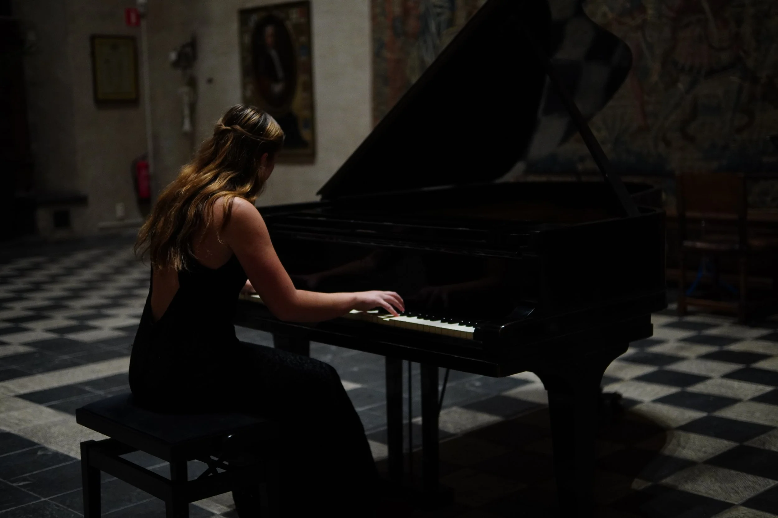 Een vrouw met lang bruin haar speelt piano in een donkerstone zaal.