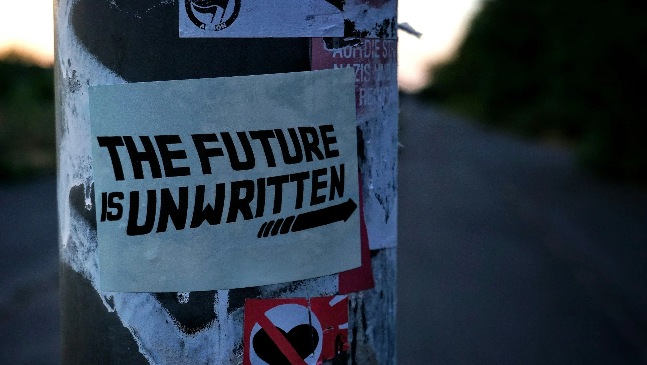 A sticker on a concrete pole that says 'The future is unwritten' with an arrow underneath. The pole is covered with other smaller stickers and posters. The background shows a road and blurred trees, indicating an outdoor setting during twilight.