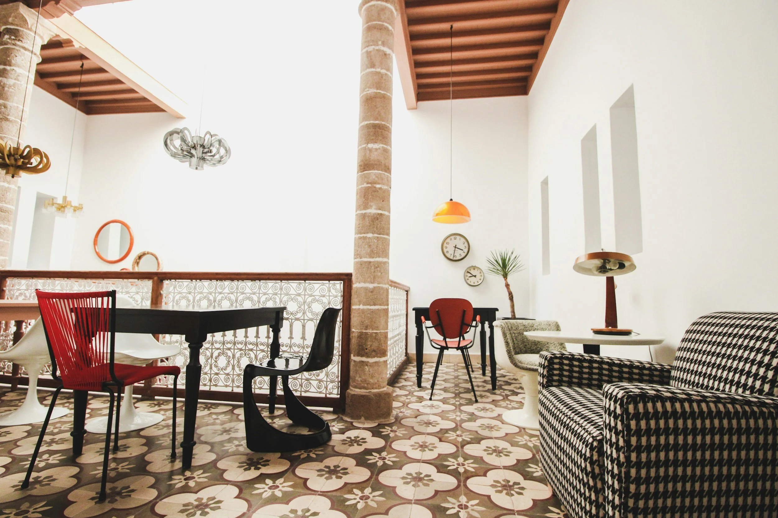 Interior of a stylish room with various chairs and tables, patterned floor tiles, a white wall with three narrow windows, and decorative wall clocks and frames.