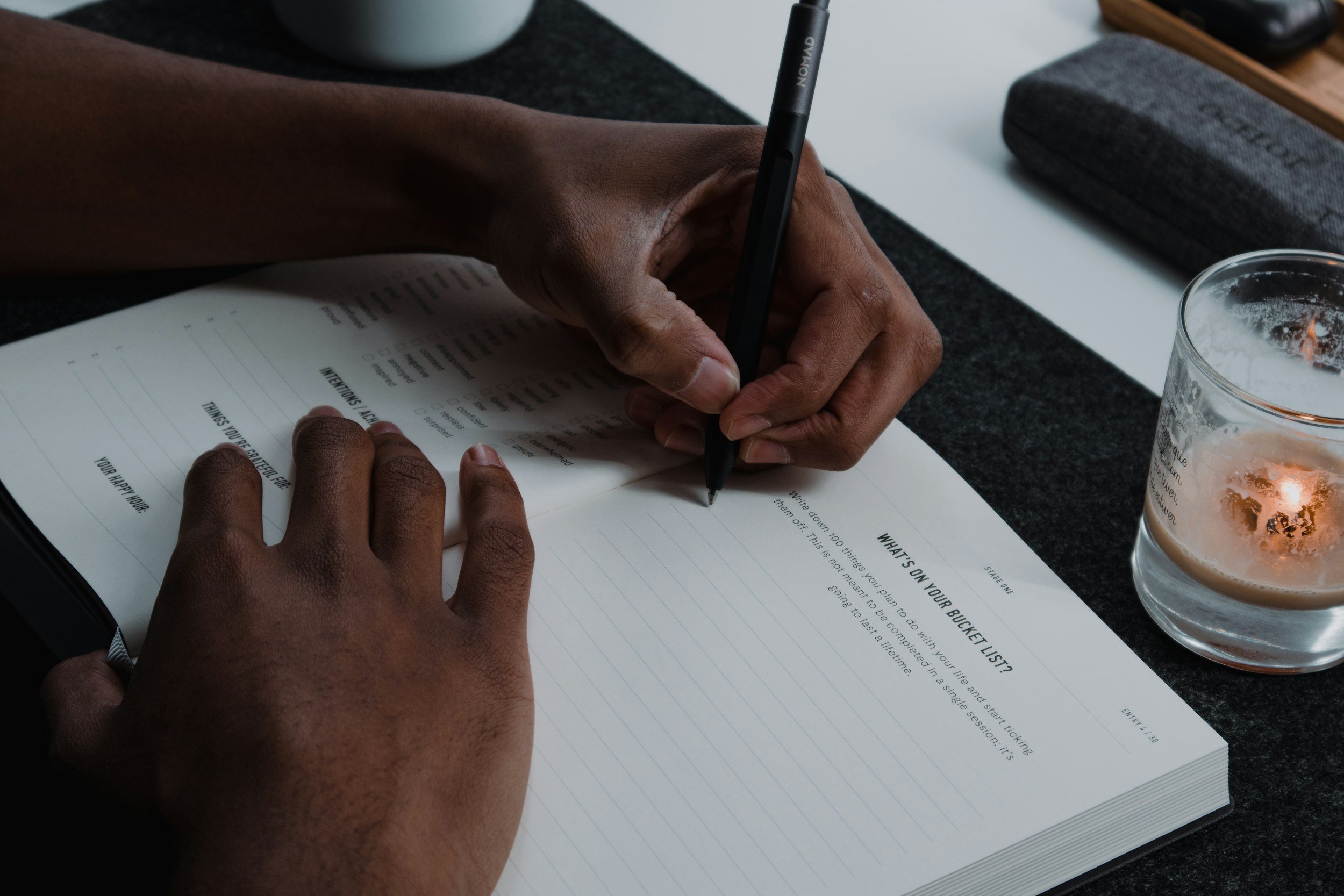 Person writing in a notebook with a black pen on a dark desk, with a glass of water and a candle nearby.