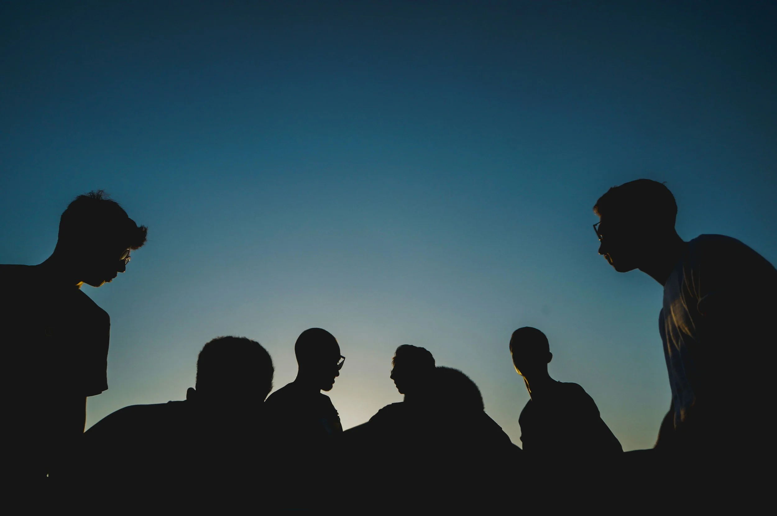Silhouettes of seven people gathered together outdoors during sunset or sunrise, with a blue sky in the background.
