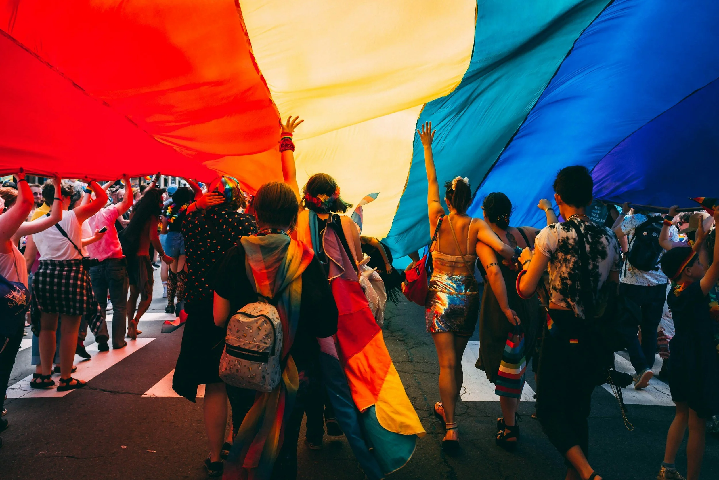 Group of diverse people participating in a pride parade, holding up a large rainbow flag.