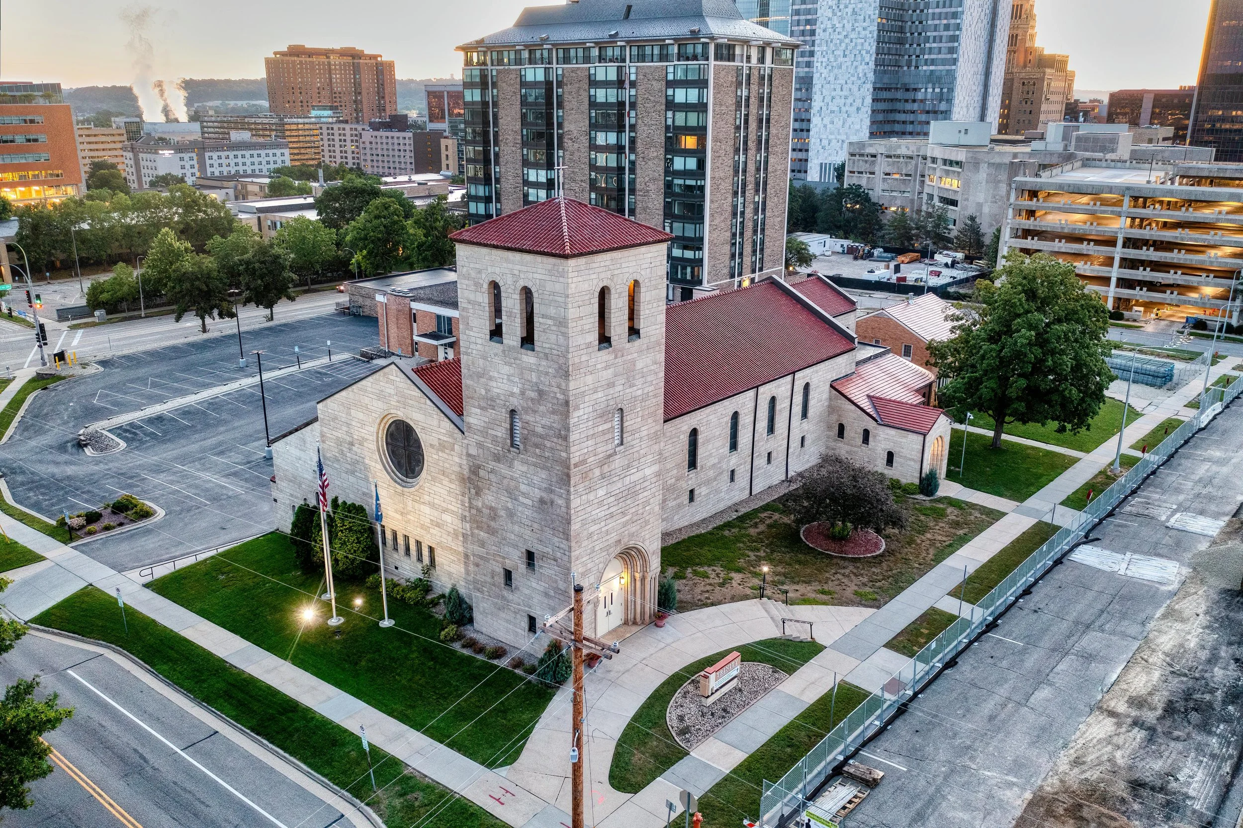 An aerial view of a historic church with a red tiled roof and stone walls, surrounded by parking lots, green lawns, and modern high-rise buildings in the background at sunset.