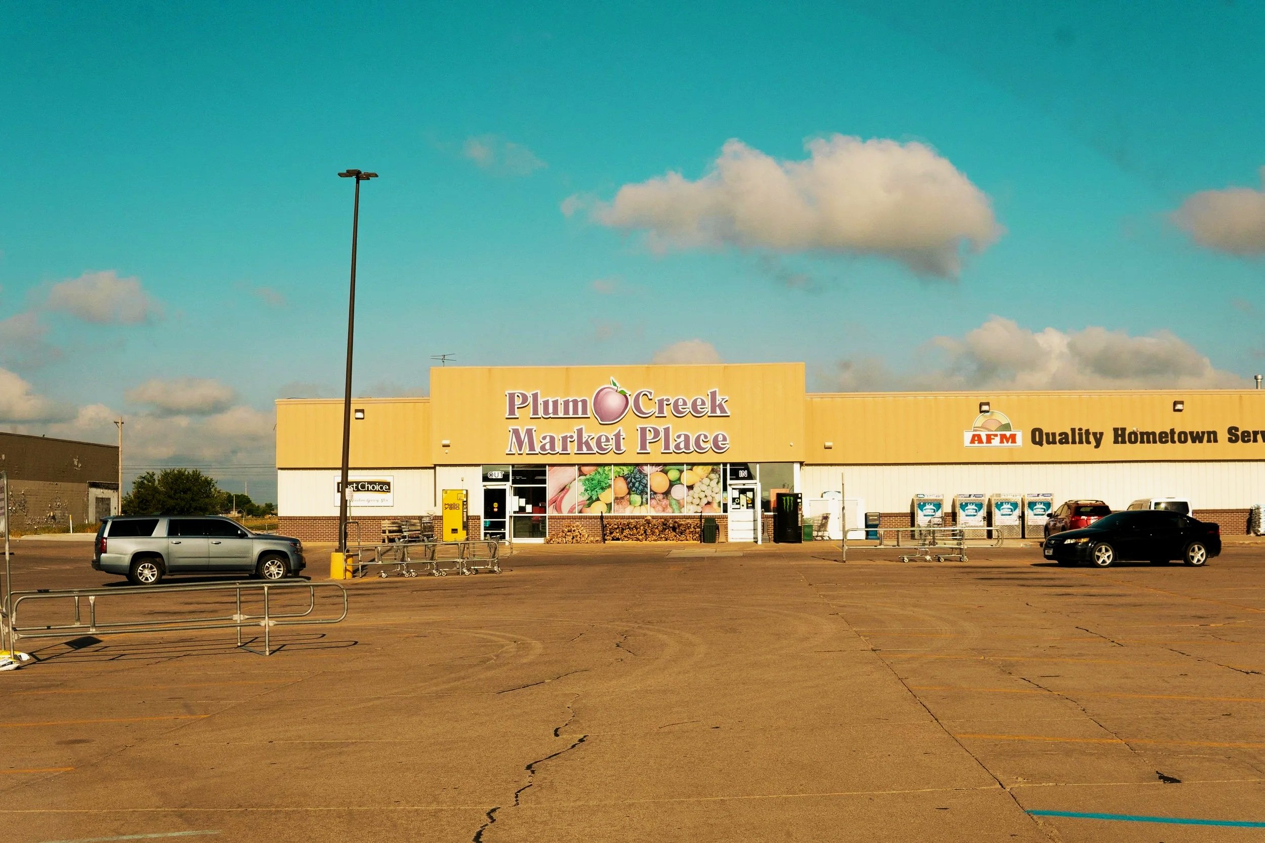 Front view of Plum Creek Market Place with parked cars and shopping carts in an empty parking lot, under a partly cloudy sky.