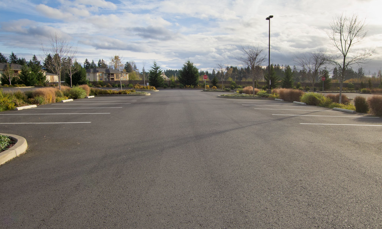 Empty parking lot with landscaped sides, no cars, trees and buildings in the background, overcast sky.