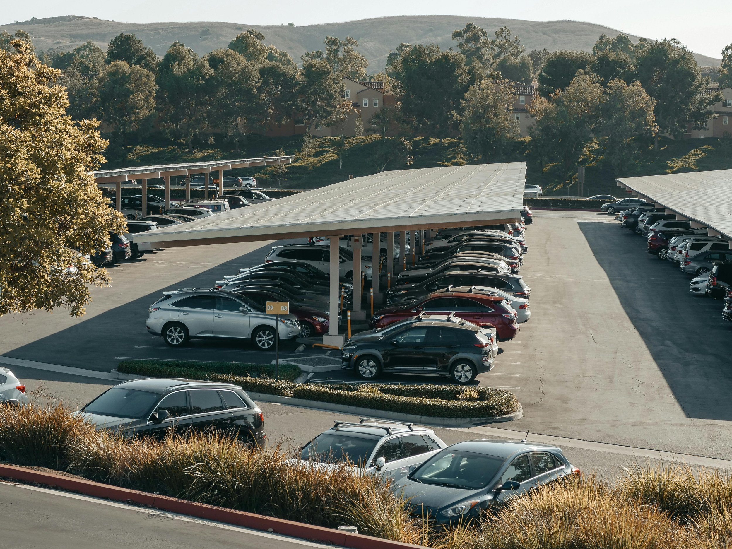 A parking lot scene with covered parking sections and multiple cars parked, with trees and hills in the background.