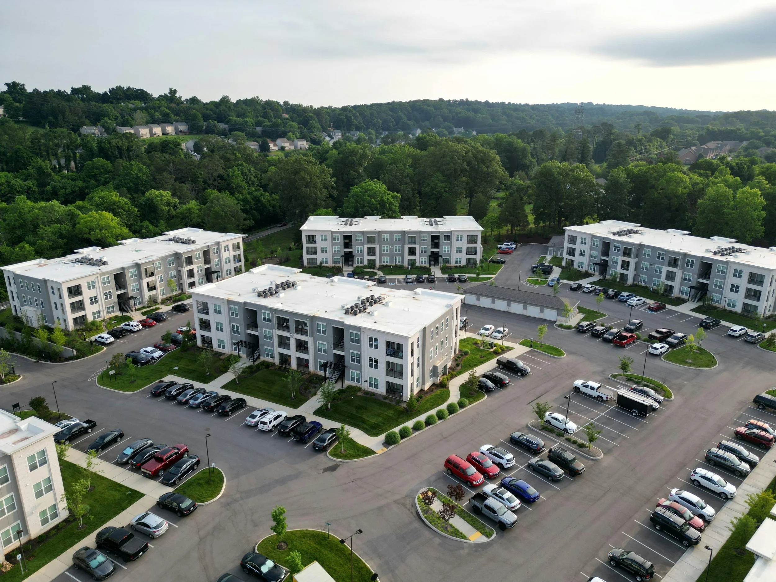 Aerial view of a residential apartment complex with multiple three-story buildings surrounded by parking lots and green landscaping, with a wooded area in the background.