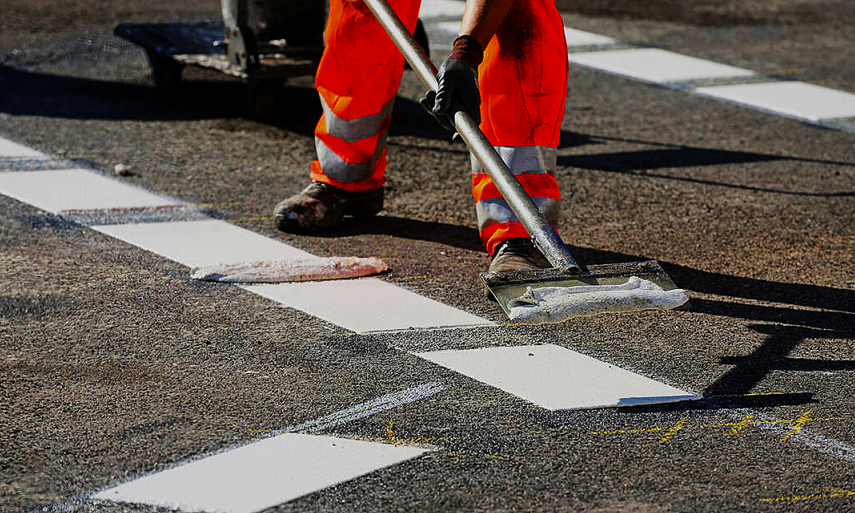 Person in orange safety clothing painting white lines on a road.