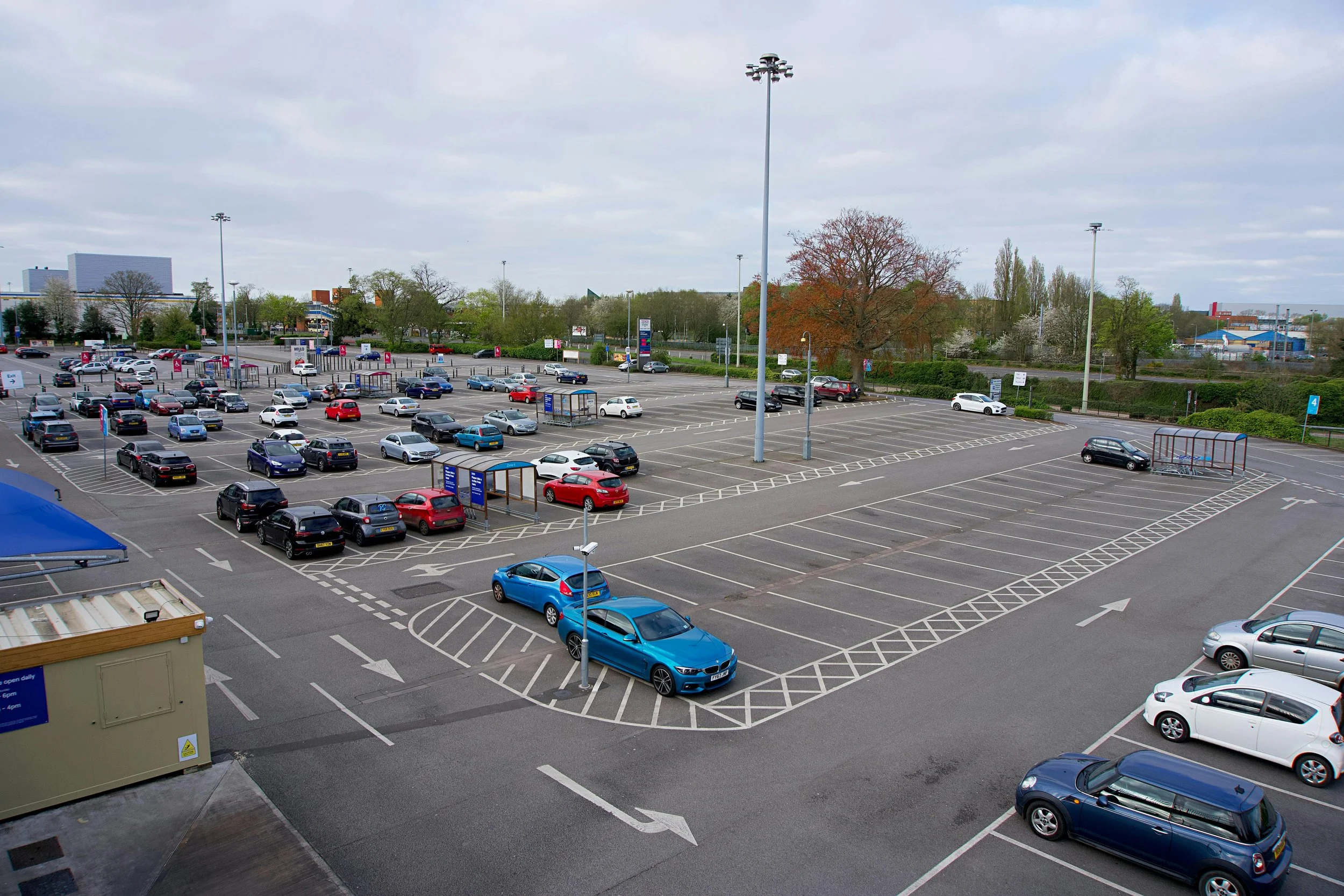 Empty parking lot with cars parked and growing trees in the background under a cloudy sky.