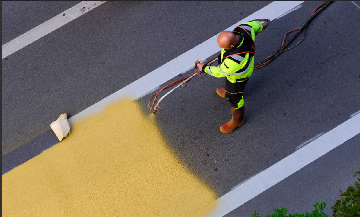 A worker in high-visibility clothing and boots is spray painting a yellow color onto a road surface using a spray gun attached to hoses.