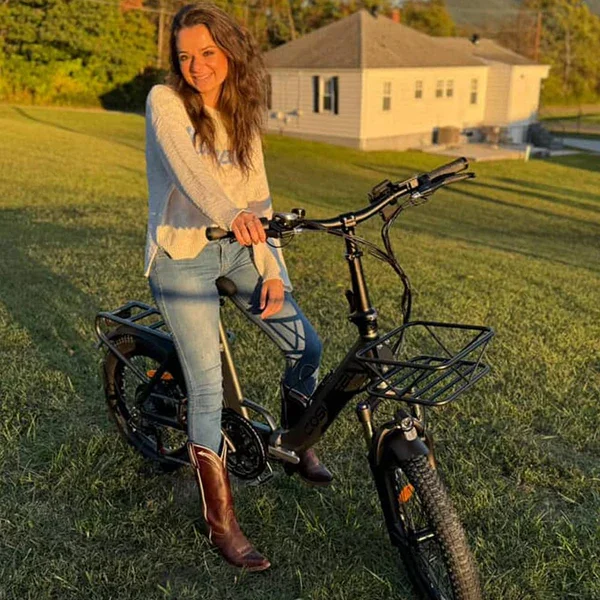 A woman with long brown hair wearing a cream sweater, jeans, and brown boots sitting on a black electric bicycle in a grassy yard during sunset.