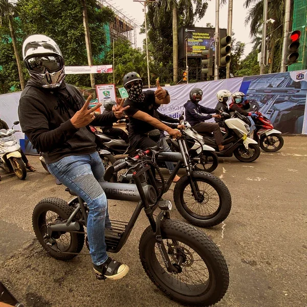 Group of motorcyclists with helmets and face masks riding or standing with bikes on a city street, with traffic lights and billboards in the background.