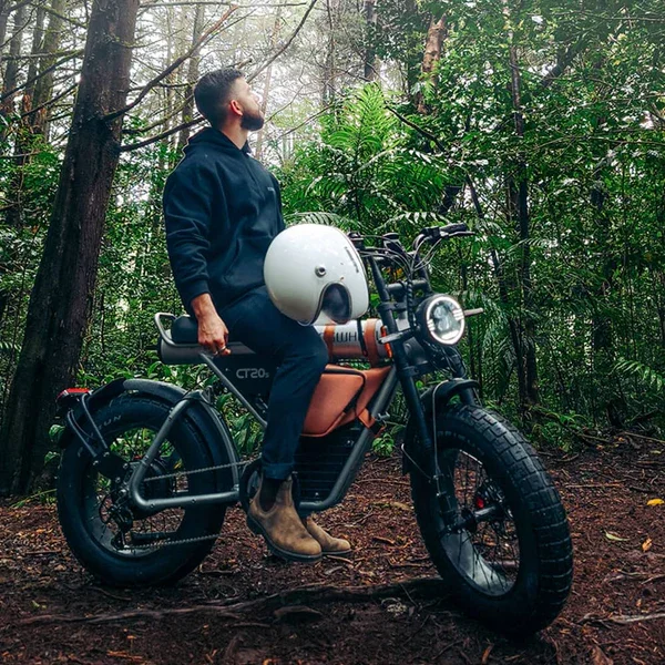 A man sitting on an electric motorcycle in a forest, holding a white helmet, wearing a black jacket and brown boots, looking up amid dense green foliage.