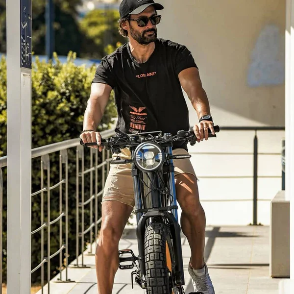A man with dark hair, beard, and sunglasses riding a black electric bicycle on a sunny sidewalk. He is wearing a black cap, black T-shirt, beige shorts, and a watch.