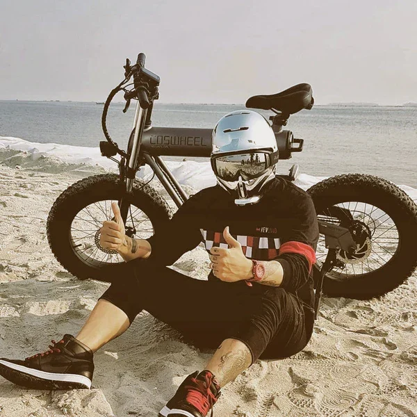 Person sitting on the sand at the beach with a helmet on, giving a thumbs-up. Behind them is an electric bicycle with fat tires, labeled 'LOSINWHEELS', and ocean in the background.