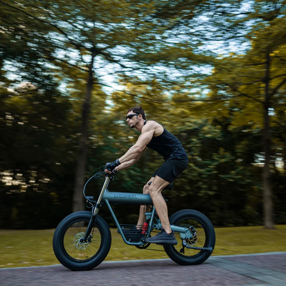 A man wearing sunglasses and athletic clothes riding an electric bike with fat tires through a park with trees and grass in the background.