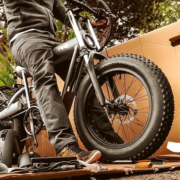 Close-up of a person standing next to a fat tire electric bicycle, with a cardboard box in the background and various tools and equipment on the floor.