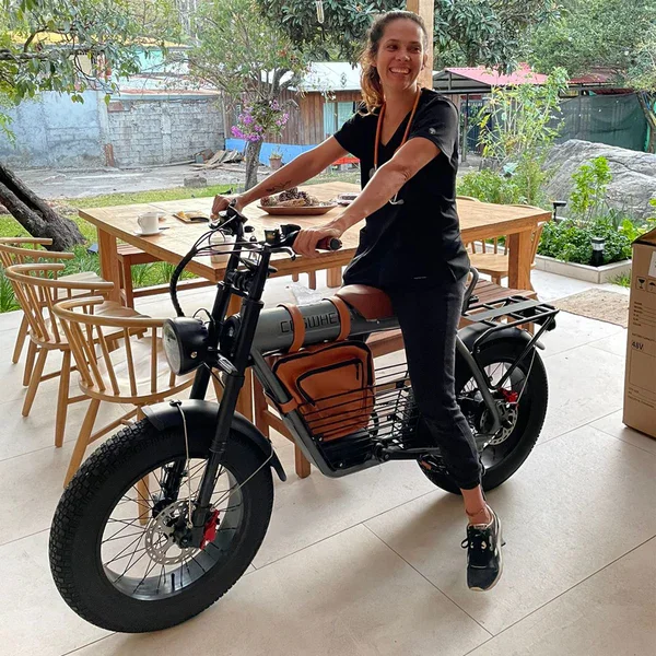 Woman in black clothes sitting on an electric bicycle, indoors with a wooden dining table and chairs, and a backyard visible through a large window.