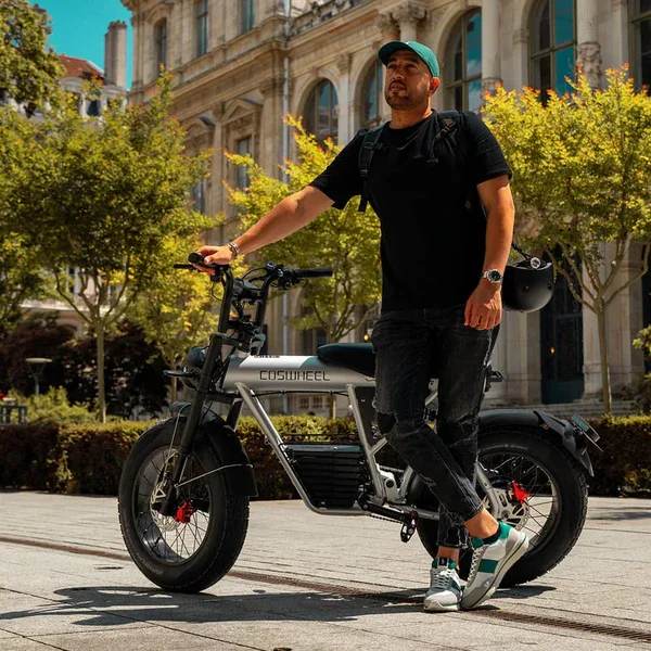 A man standing next to an electric bicycle outdoors in an urban area with trees and historic buildings in the background.