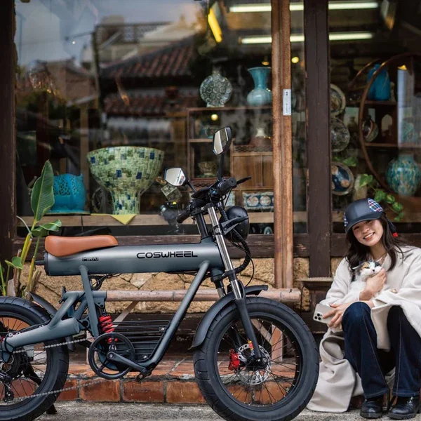 A woman wearing a black cap, white coat, and jeans sitting on the sidewalk next to a gray electric bike with a brown seat, holding a small dog and smiling.