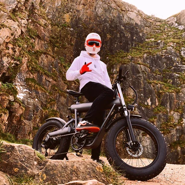 Person wearing a white helmet with red accents, white hoodie, black pants, and red gloves, sitting on a black mountain bike in front of a rocky hillside, making a rock and roll hand gesture.