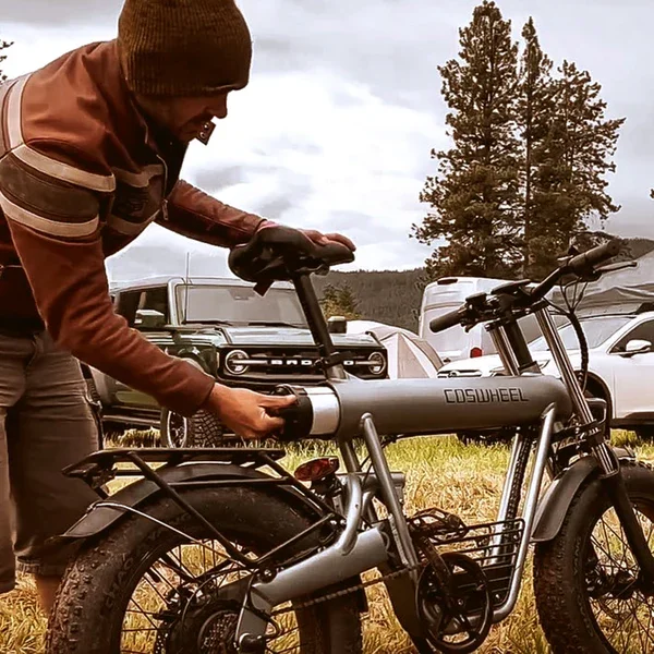 A man in a brown jacket and beanie working on a gray e-bike labeled COSWHEEL outdoors in a grassy area with parked cars and trees in the background.