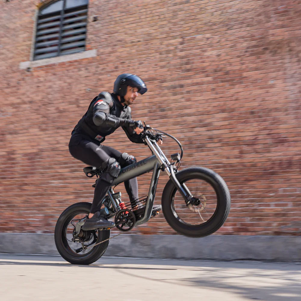 Man wearing helmet and protective gear riding a black electric bicycle with front wheel lifted off the ground in an urban setting with a brick wall background.