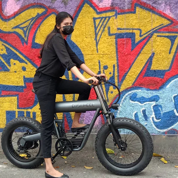 Woman wearing a black mask and black clothing, sitting on a black fat-tire electric bicycle in front of a colorful graffiti wall.