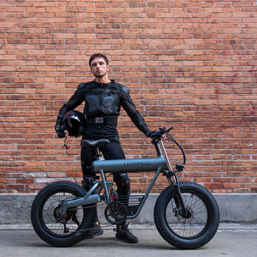 A man in black riding gear standing next to an electric mountain bike against a brick wall, holding a helmet.