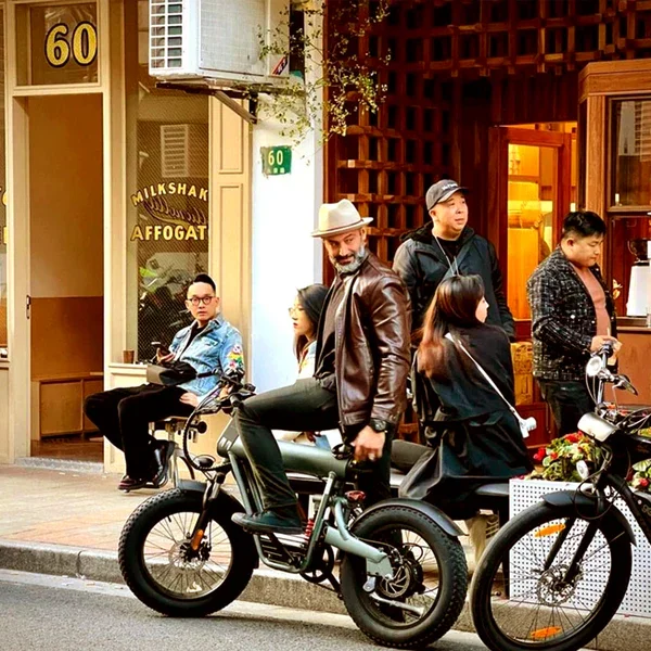 Group of five people waiting outside a cafe with a motorcycle parked nearby, in an urban street scene.
