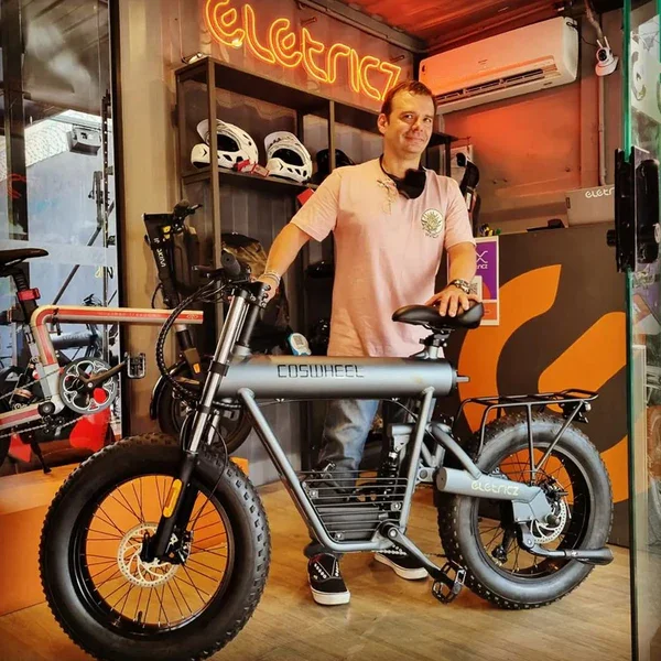 Man standing inside a store with an electric bicycle, helmet display, and neon sign reading 'ELECTRIC' in the background.