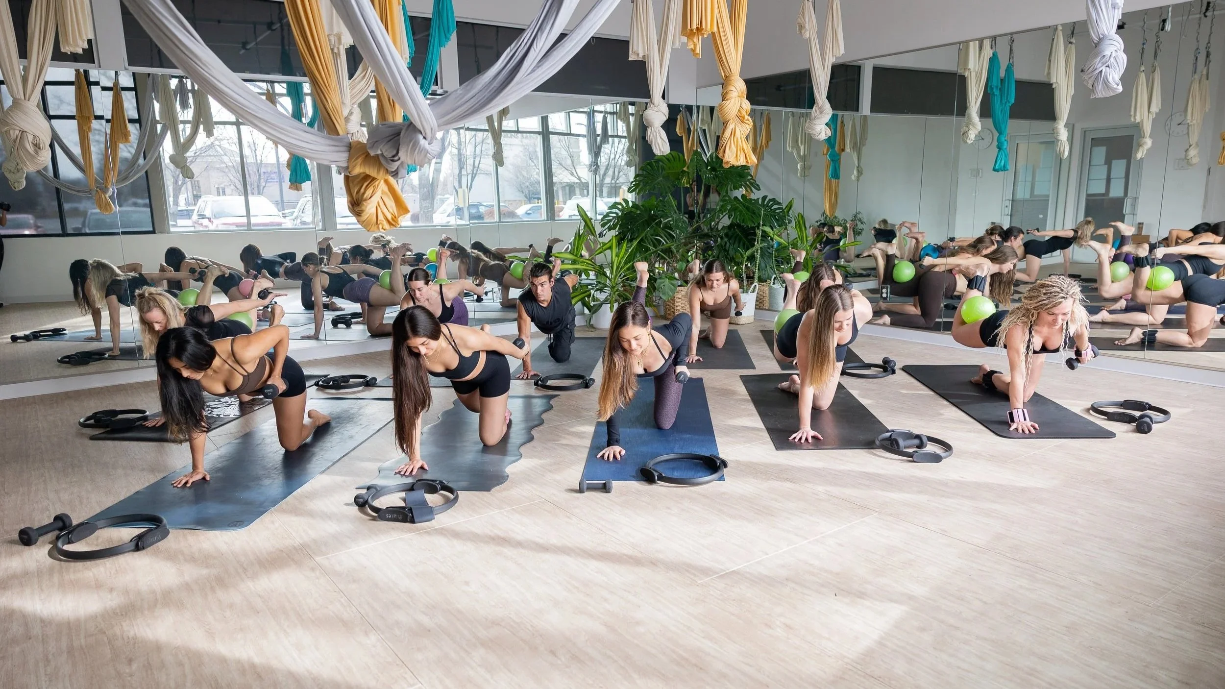 Women participating in a yoga or fitness class in a studio with mirrors, plants, and aerial fabric installations overhead. They are on yoga mats, doing various stretches and poses with dumbbells and resistance rings.