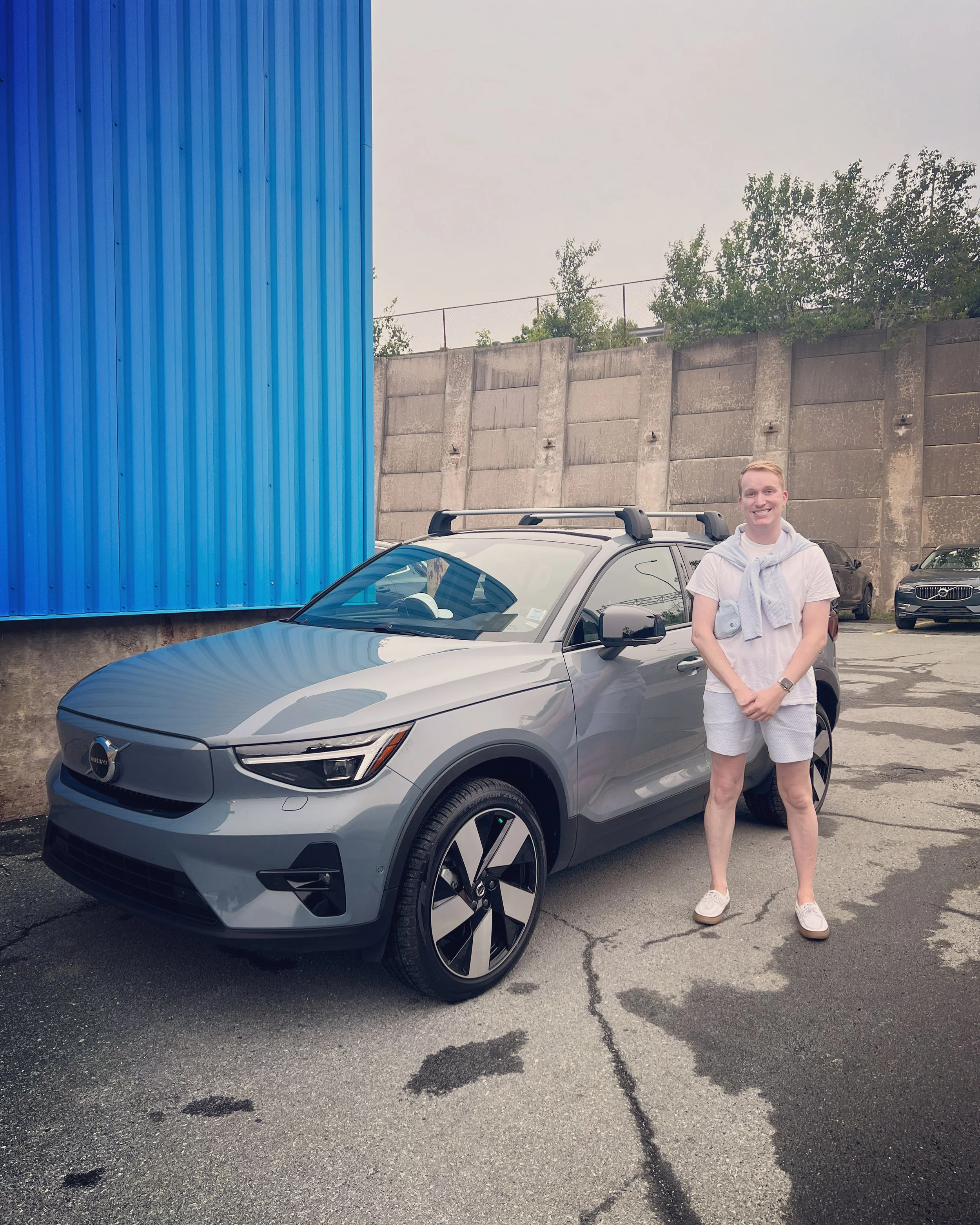 A smiling young man stands in front of a gray car in a parking lot, with a blue building and a concrete wall with trees in the background.