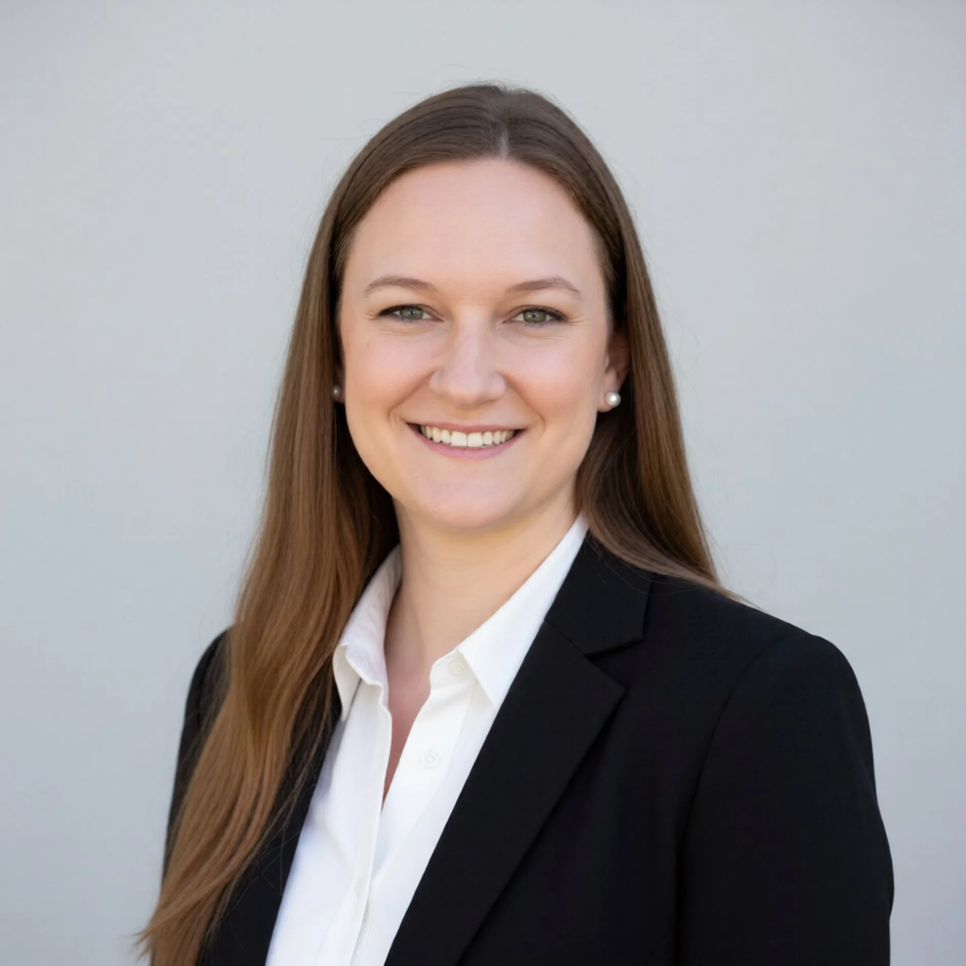 A professional woman with long brown hair, wearing pearl earrings, a white blouse, and a black blazer, smiling against a plain light gray background.
