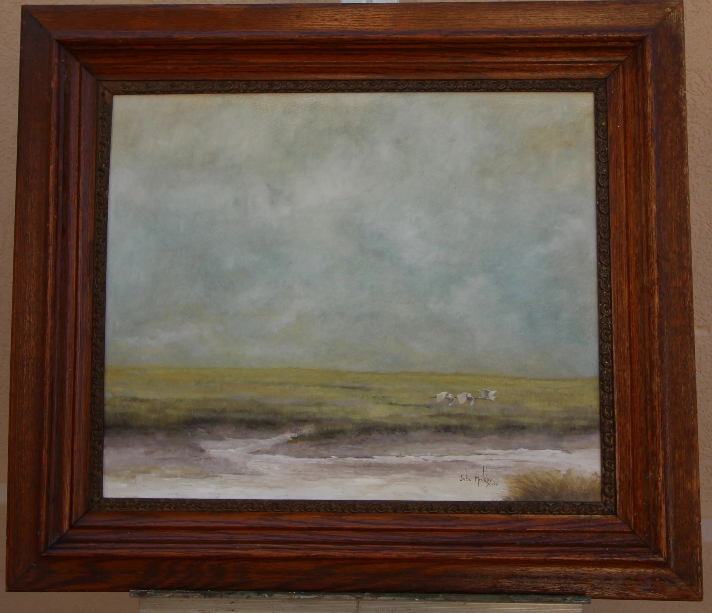 A framed landscape painting of a cloudy sky, green field, and a few white birds flying low over the ground.